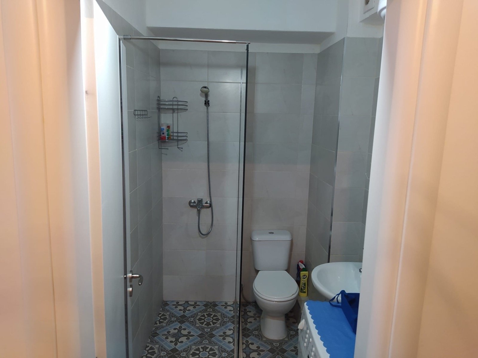 Modern bathroom with glass shower enclosure, white toilet, decorative floor tiles with geometric pattern, metal shower fixtures and shelves